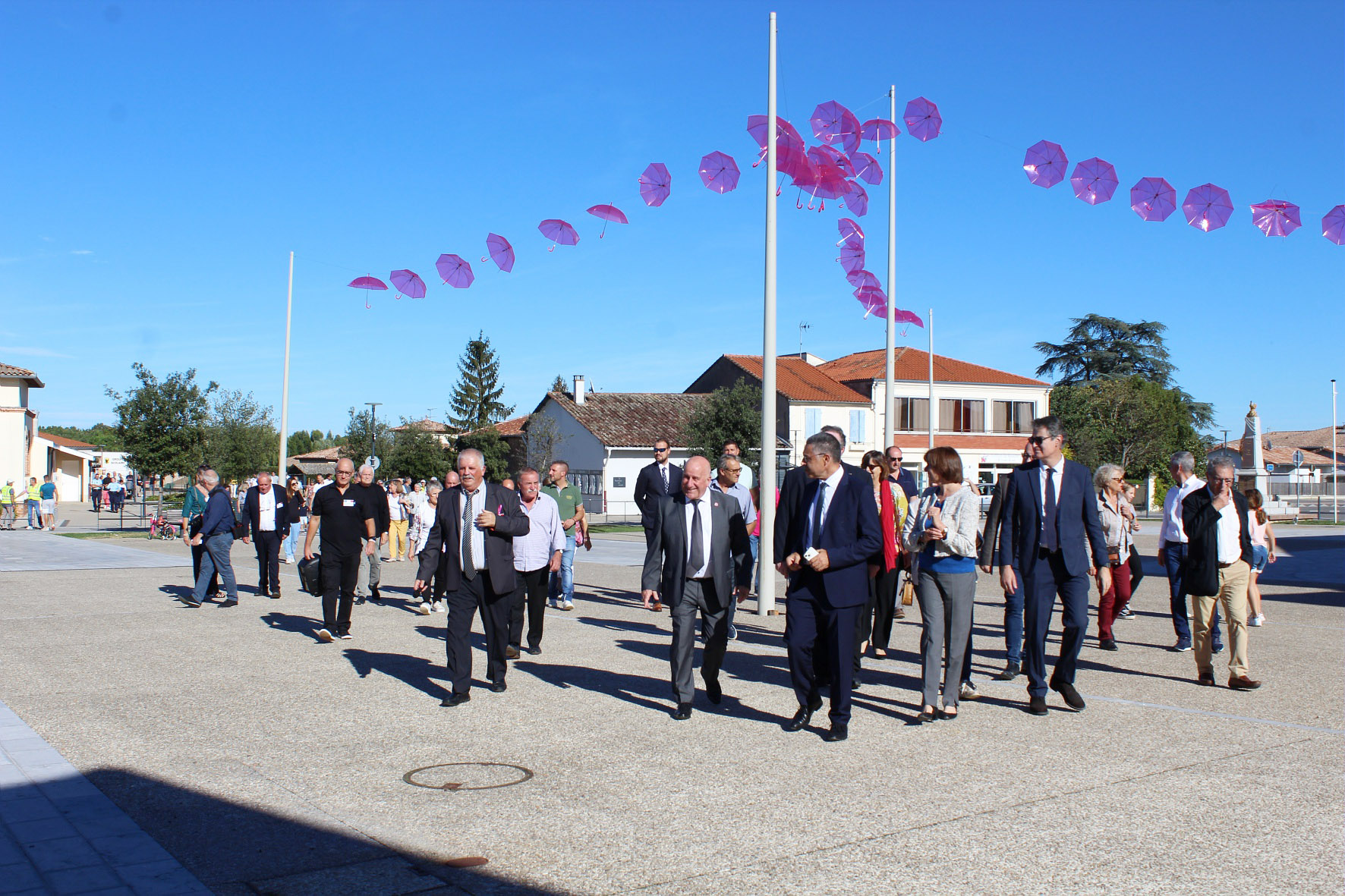 Les élus et le président qui marchent sur la place du village décorée avec des parapluies roses 