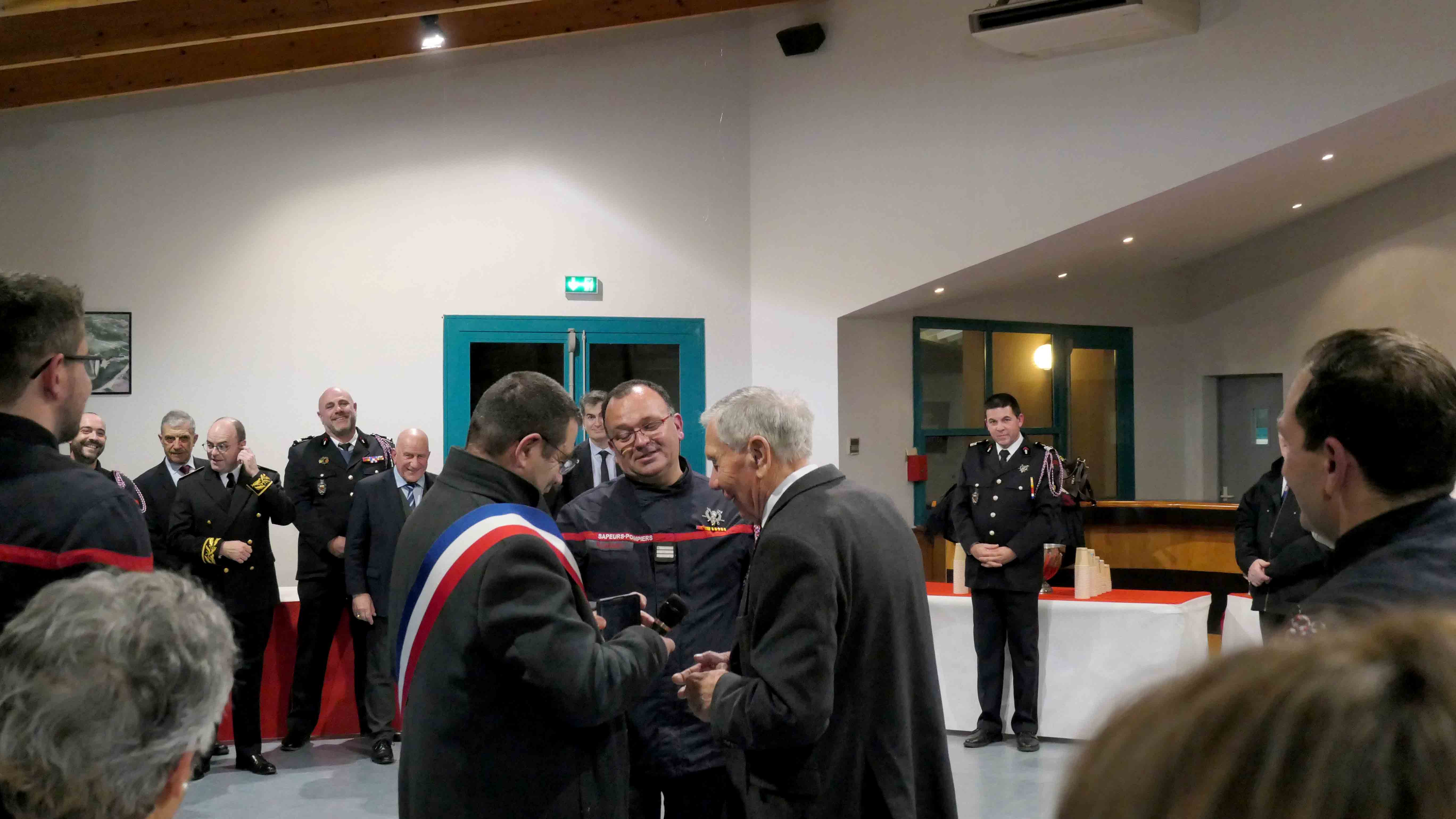 M.Le maire de Montpezat-de-Quercy qui remet une médaille à un pompier dans une salle en présence de nombreux sapeurs-pompiers et de Michel Weill