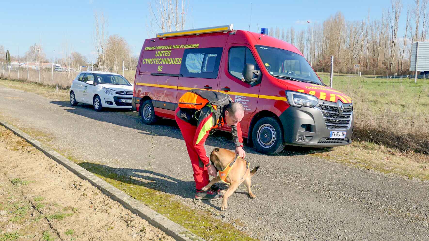 Un maître-chien et un chien sauveteur, lors d'un entraînement, et un camion de pompier pour le transport de chiens sauveteurs en arrière plan
