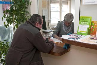 Aide à l'hébergement séniors Une personne âgée s’appuie sur un comptoir à l’accueil pendant qu’une autre, derrière le guichet, remplit un formulaire dans son bureau