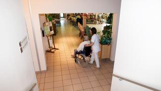 On peut voir une aide soignante entrain d'aider une sénior en chaise roulante 