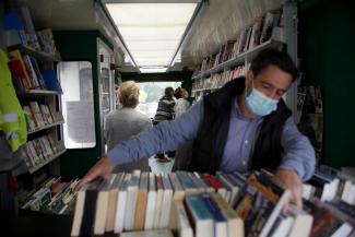 L'intérieur du bibliobus départemental Un homme en train de chercher des livres dans une caisse (premier plan) et des usagers qui déambulent dans le bibliobus
