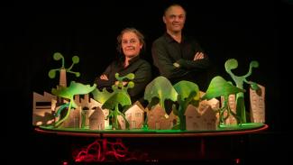 Spectacle "L'Arbre" à l'abbaye de Belleperche Deux personnes qui croisent les bras en regardant l'objectif avec devant eux une maquette avec des maisons et des arbres en cartons