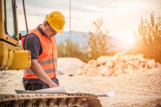 Visuel : chantier Un homme en tenue de chantier en train de lire un plan posé sur des chenilles de tractopelle
