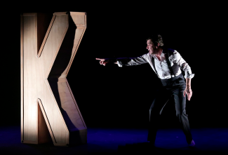 Pièce de théâtre "Le K" de Dino Buzzati à l'espace des Augustins Un homme montrant du doigt une lettre K en bois géante