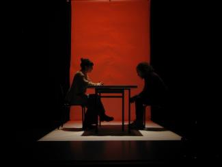 Spectacle théâtral "nos rêvoltes" joué à l'espace des Augustins Un homme et une femme assis dans le noir à table avec à l'arrière un fond rouge